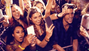 A lively crowd of young adults at a concert, smiling and cheering near the front. Several people hold up smartphones to take photos or videos, and colorful lights create an energetic atmosphere.