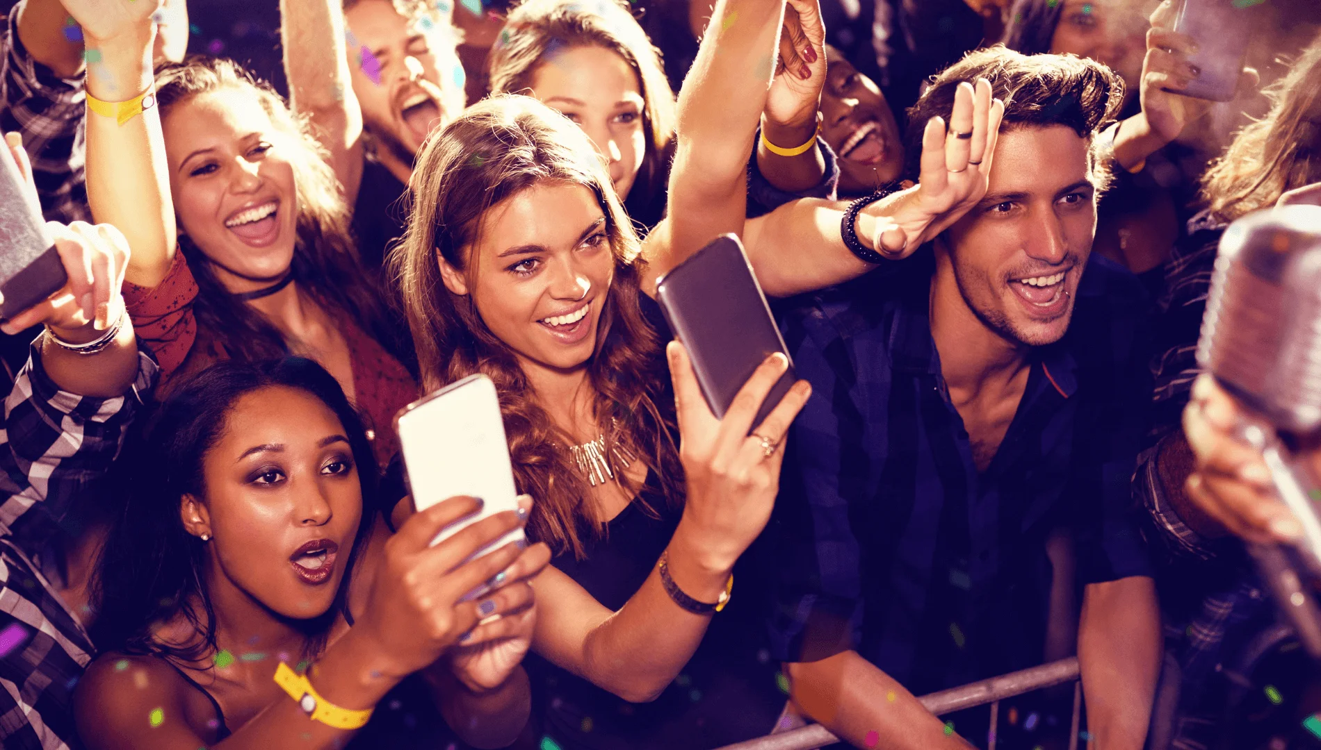 A lively crowd of young adults at a concert, smiling and cheering near the front. Several people hold up smartphones to take photos or videos, and colorful lights create an energetic atmosphere.