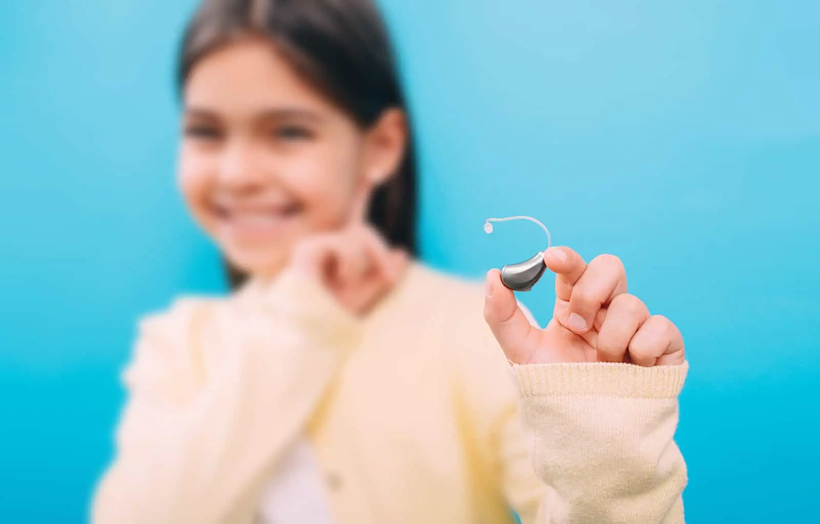 A smiling child in a yellow sweater holds up a hearing aid in front of a blue background, with the focus on the hearing aid and the child slightly blurred.