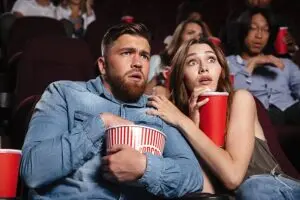 A man and woman sit closely together in a movie theater, both looking surprised and slightly scared. The man holds a tub of popcorn while the woman clings to his arm. Other people with drinks sit behind them.