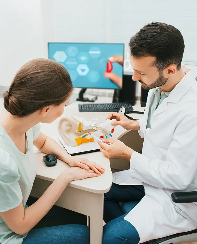A doctor in a white coat shows a female patient an anatomical model of the ear while explaining hearing-related information in a medical office. A computer screen with medical icons is visible in the background.