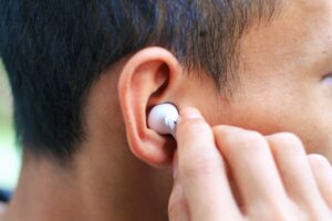 A close-up of a person inserting a white wireless earbud into their ear, with their hand holding the earbud and part of their face and hair visible.