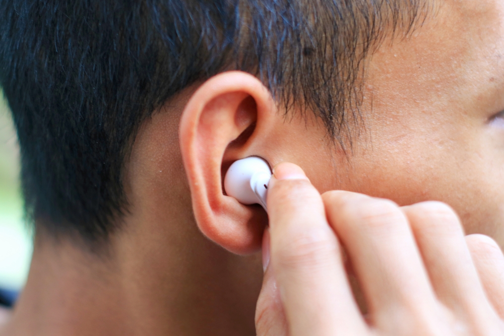 A close-up of a person inserting a white wireless earbud into their ear, with their hand holding the earbud and part of their face and hair visible.