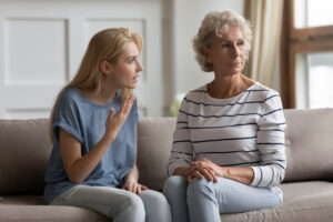 A younger woman is talking emotionally to an older woman who looks away with a serious expression. They are sitting together on a couch in a bright living room.