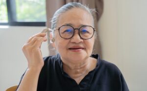 An older woman with gray hair and glasses smiles while holding a hearing aid to her ear. She wears a black top and sits indoors near a window with natural light coming in.