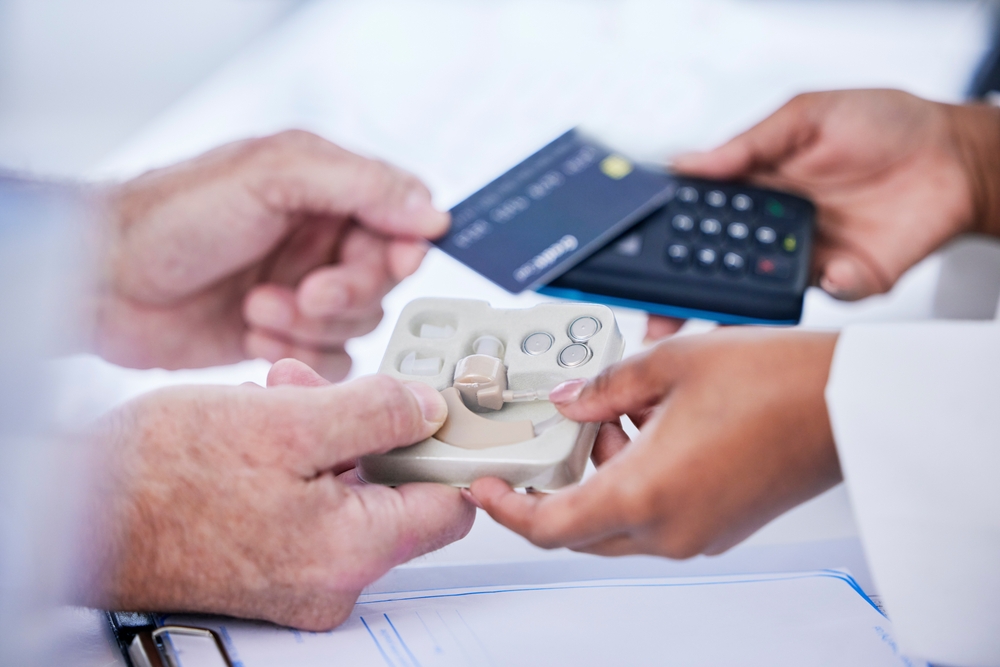 A person pays for a hearing aid device using a credit card and a card reader held by another person, with paperwork visible on the table below.