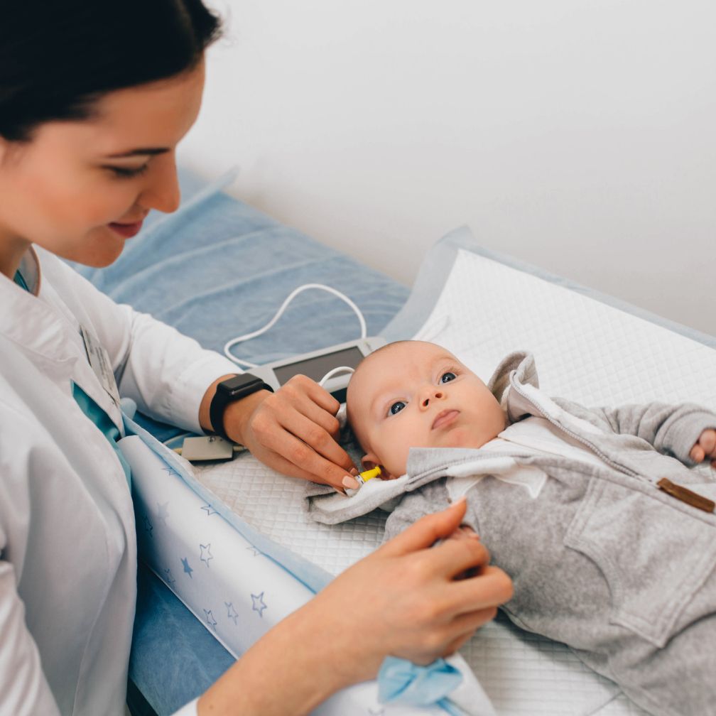 A healthcare professional tests a babys hearing using specialized equipment while the baby lies on an examination table in a clinic.