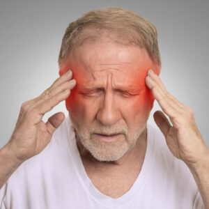 Older man with closed eyes, holding his temples with both hands and appearing to be in pain. Red shading highlights his temples, indicating a headache or migraine. He wears a white shirt and has gray hair and beard.