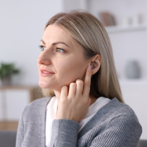 A woman with blonde hair touches her ear and looks to the side, appearing thoughtful. She wears a gray cardigan and sits indoors with a blurred background.