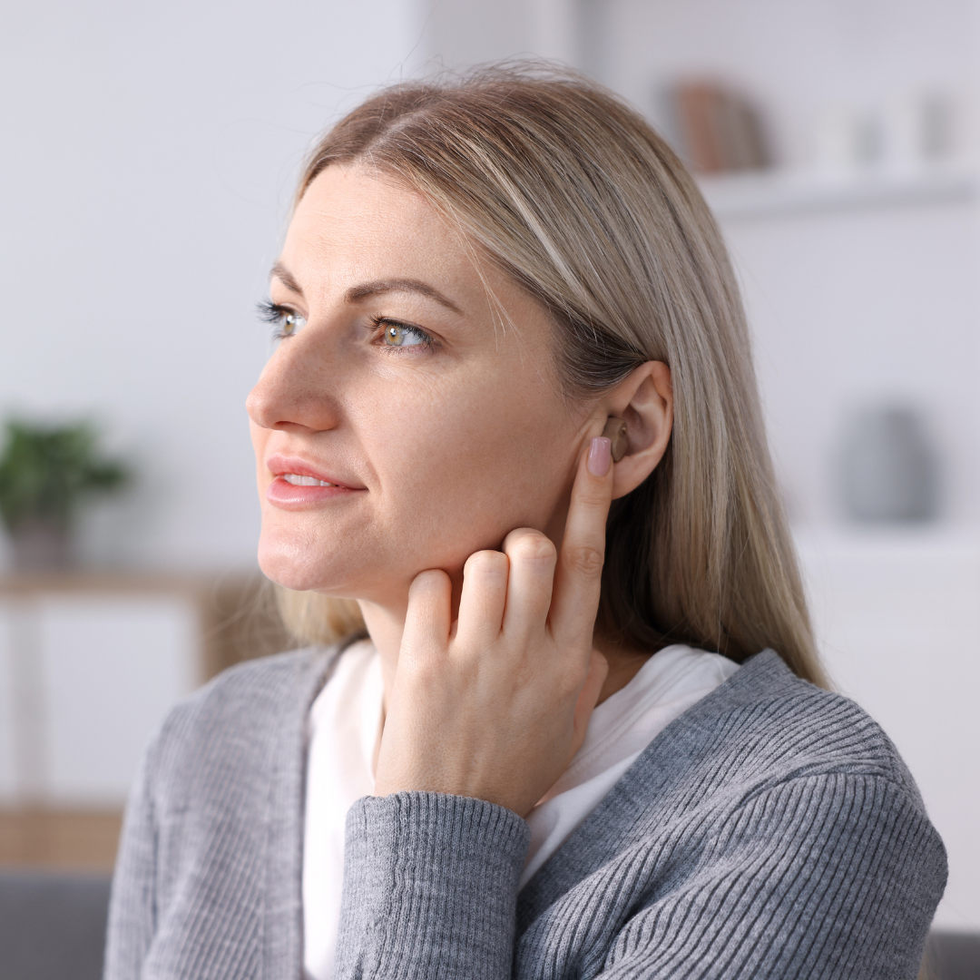 A woman with blonde hair touches her ear and looks to the side, appearing thoughtful. She wears a gray cardigan and sits indoors with a blurred background.